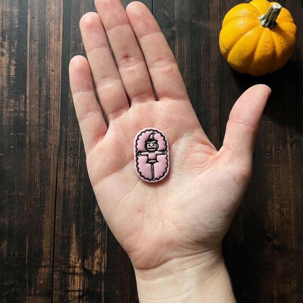 Finished pink pumpkin scarecrow clay piece resting in an open hand for scale, showing the jack-o-lantern face, pointed hat, outstretched arms, and rounded border.