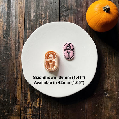 Orange pumpkin scarecrow polymer clay cutter beside a finished pink clay example with black detailing, displayed on a white round surface with a pumpkin in the background.