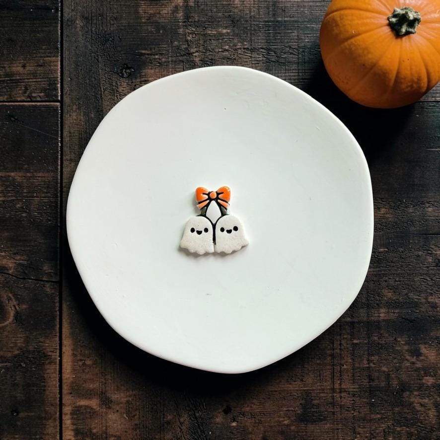 Finished ghost cherries clay piece centered on a white plate, showing two white ghosts with tiny black faces connected beneath an orange bow.
