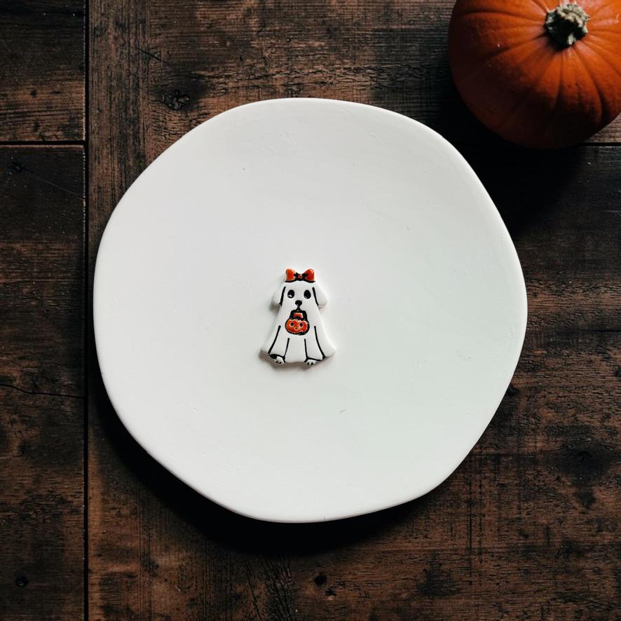 Finished white Halloween dog clay piece centered on a white plate showing a puppy in a ghost costume with an orange bow and jack-o-lantern treat bucket.