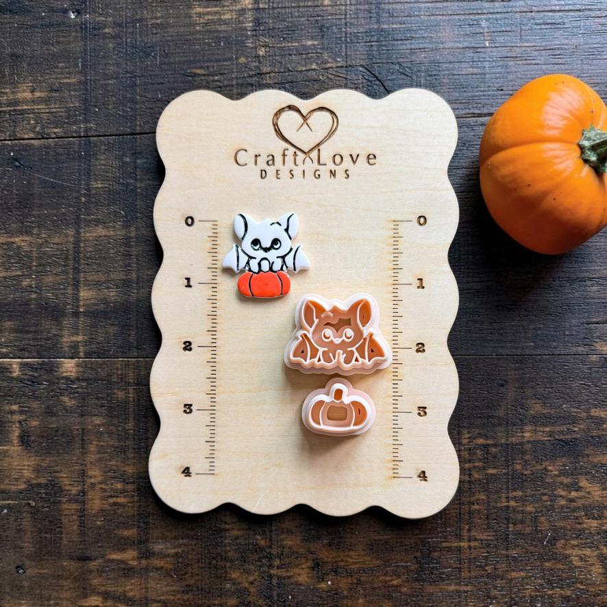 Finished white and orange bat on pumpkin clay piece beside the matching peach bat and separate pumpkin designs on a Craft Love Designs wooden measuring board.