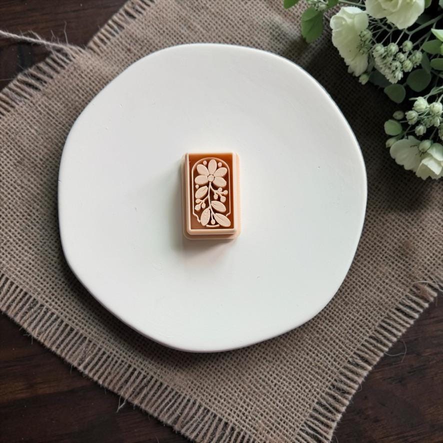 Peach rectangular polymer clay cutter centered on a white plate, featuring an embossed flower, leafy branch, and small botanical details.