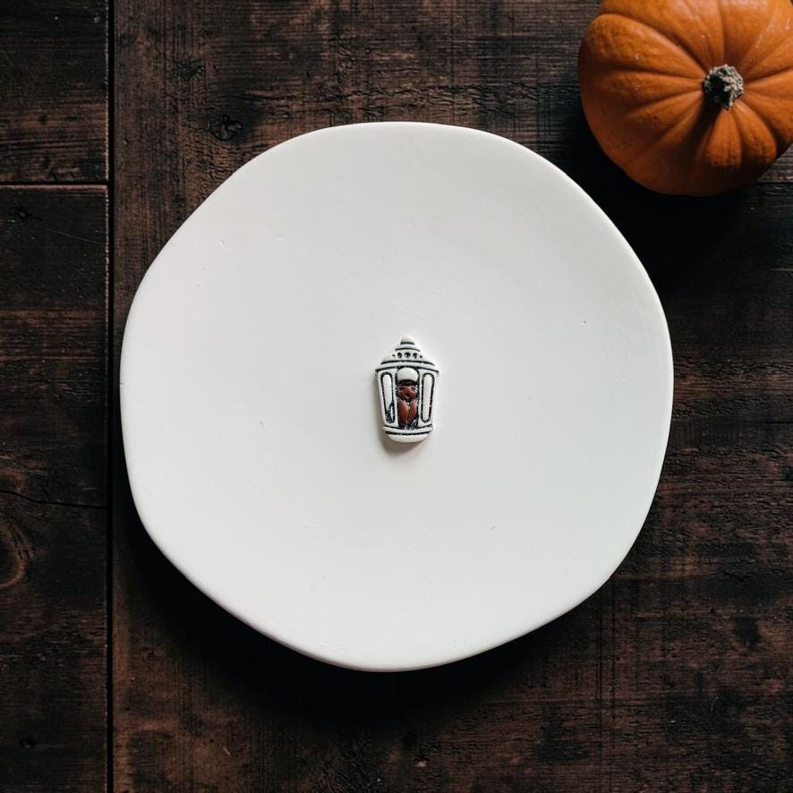 Finished white and black miniature lantern clay piece featuring a bat inside, displayed on a white plate with a pumpkin nearby.