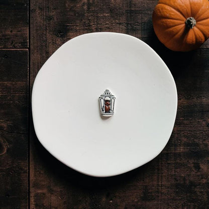 Finished white and black miniature lantern clay piece featuring a bat inside, displayed on a white plate with a pumpkin nearby.