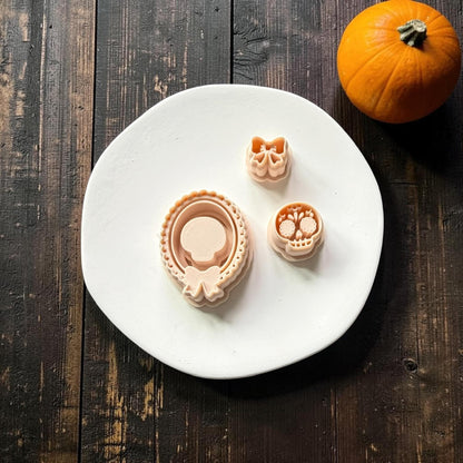 Peach three-piece polymer clay cutter set arranged on a white round surface, including an ornate oval frame with bow, decorative sugar skull, and separate bow, with an orange pumpkin in the background.