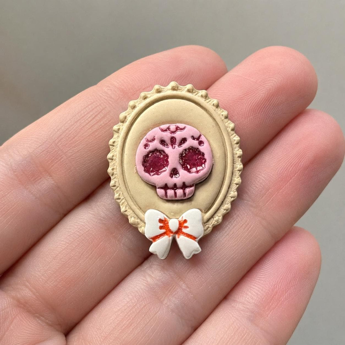 Finished 50mm sugar skull cameo held in a hand for scale, showing the pink decorative skull inside a cream ornate oval frame with a white and orange bow.