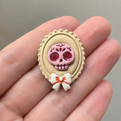 Finished 50mm sugar skull cameo held in a hand for scale, showing the pink decorative skull inside a cream ornate oval frame with a white and orange bow.