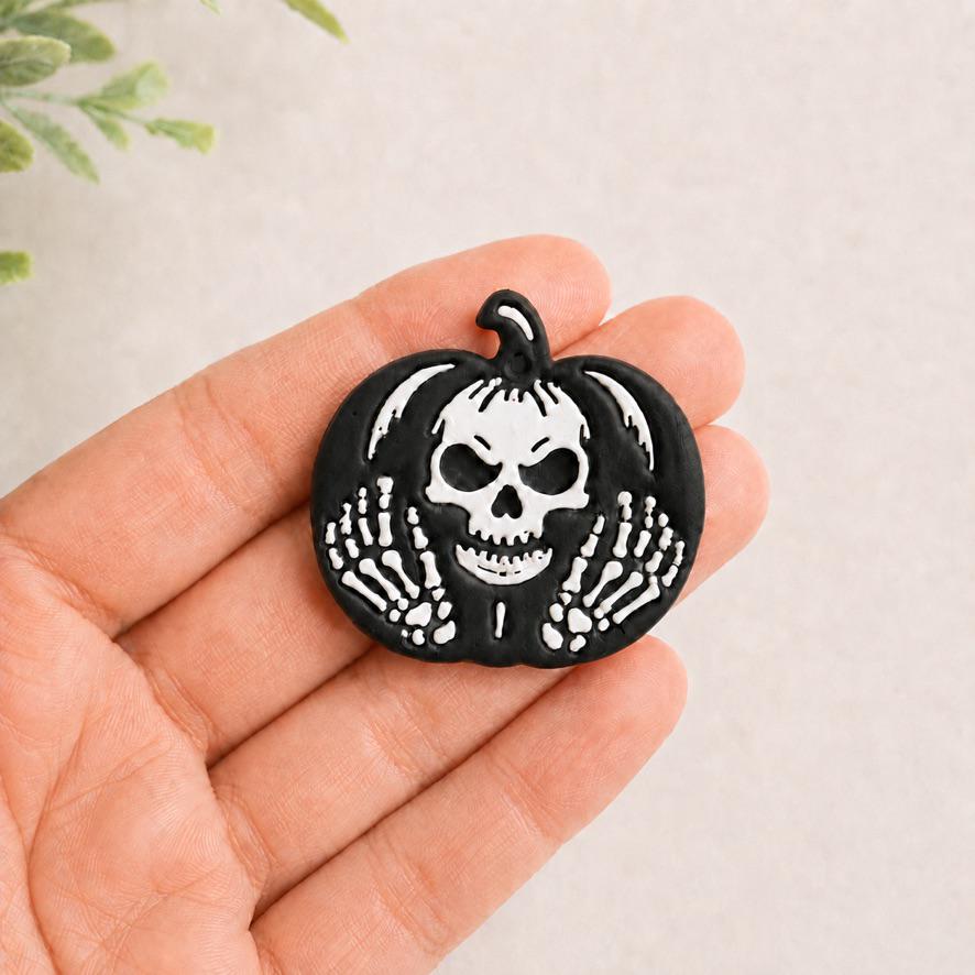 Finished black and white skeleton pumpkin clay piece held in a hand for scale, featuring a central skull, pumpkin stem, and skeletal hands on both sides.