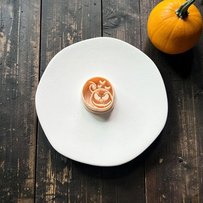 Peach pumpkin face polymer clay cutter centered on a white plate, featuring a large spooky grin, expressive eyes, curled stem, and three bats.