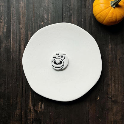 Finished white clay pumpkin face piece with black details centered on a white plate, showing the grinning face, curved pumpkin outline, curled stem, and three bats.