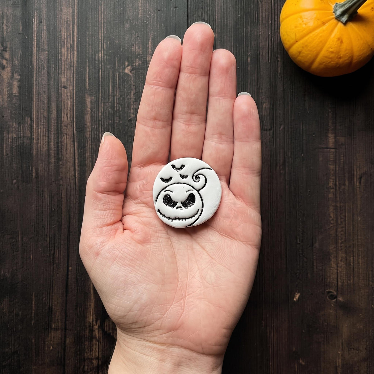 Finished white and black spooky pumpkin face clay piece displayed in an open hand for scale, featuring a large creepy smile, curled stem, and three flying bats.