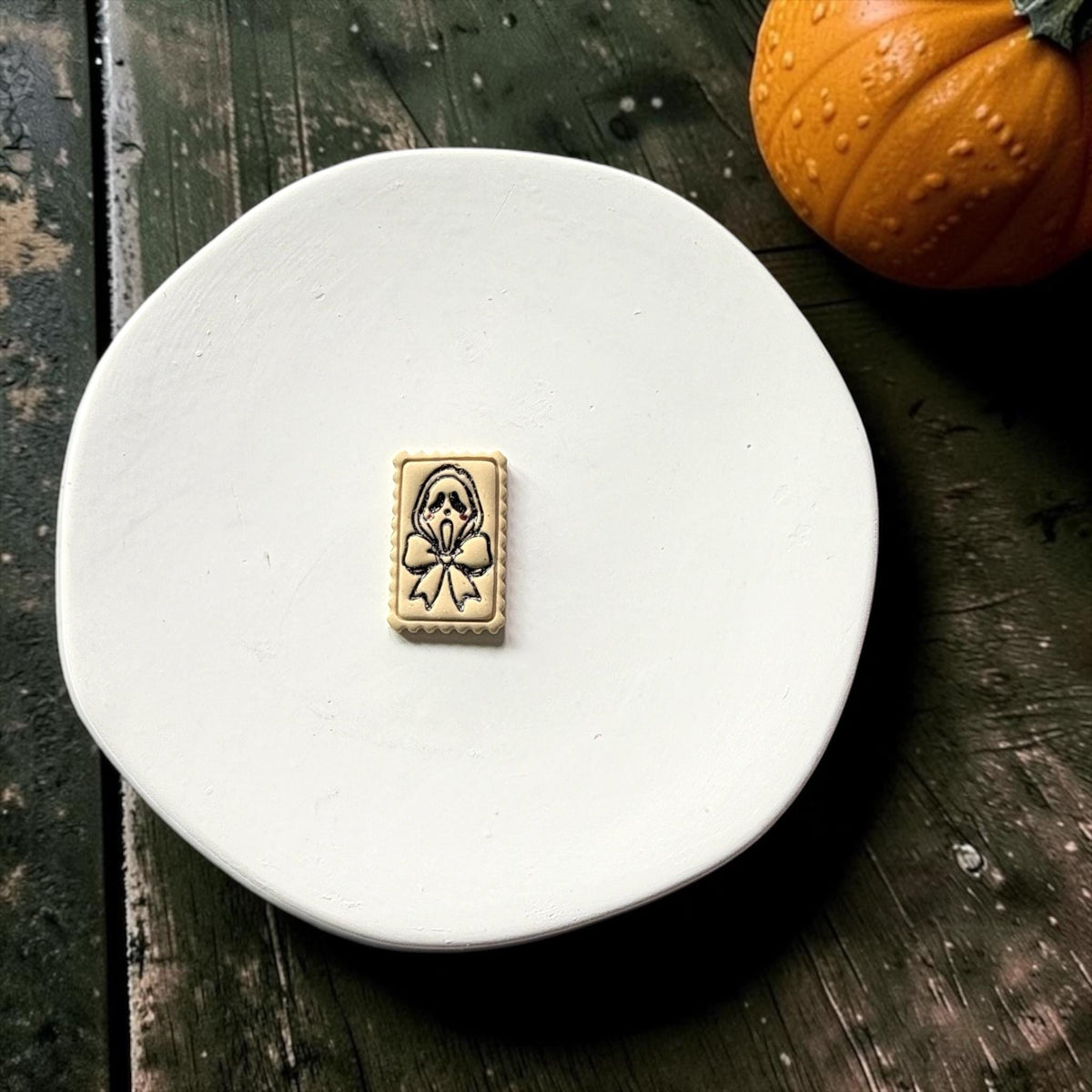 Finished cream rectangular clay piece centered on a white plate, featuring a black outlined spooky ghost face, large bow, and scalloped frame with a pumpkin in the background.