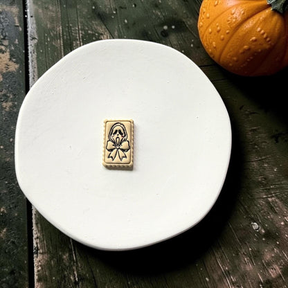 Finished cream rectangular clay piece centered on a white plate, featuring a black outlined spooky ghost face, large bow, and scalloped frame with a pumpkin in the background.
