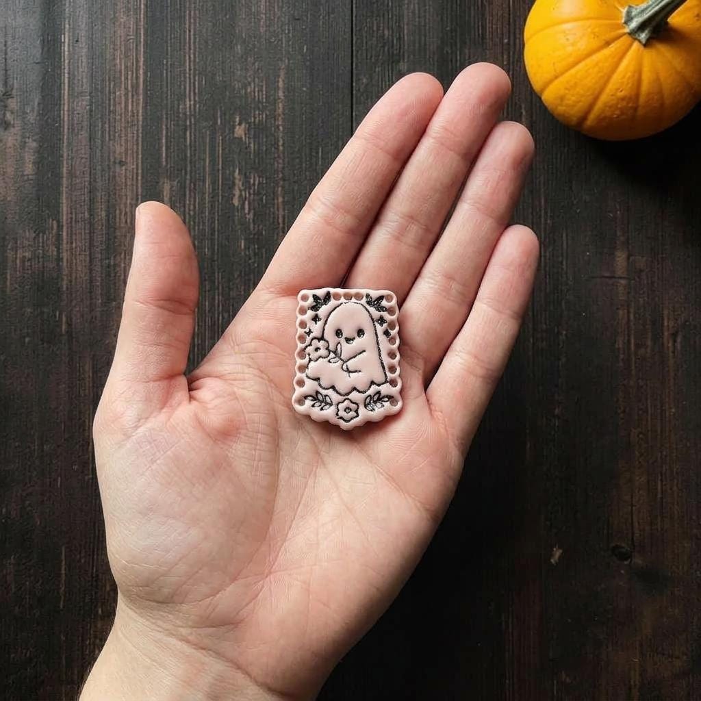 Finished pale pink floral ghost clay piece resting in an open hand for scale, showing the ghost, bouquet, flowers, leaves, bats, stars, and decorative border.