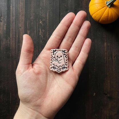 Finished pale pink floral ghost clay piece resting in an open hand for scale, showing the ghost, bouquet, flowers, leaves, bats, stars, and decorative border.