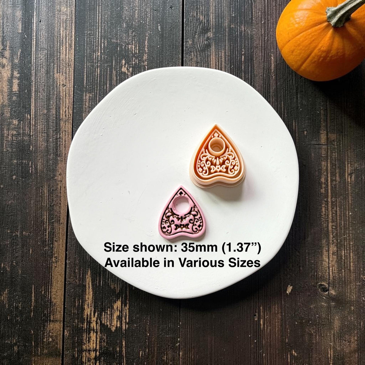Peach planchette polymer clay cutter beside a finished pink example on a white plate, showing an ornate crescent moon and scrollwork design with a circular opening. Text states 35mm or 1.37 inches and various sizes available.