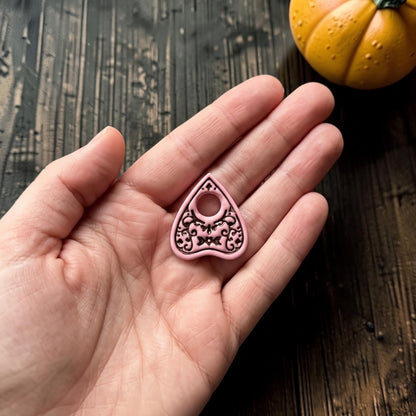 Finished pink and black planchette-shaped clay piece resting in an open hand for scale, highlighting the circular opening, crescent moon motif, pointed silhouette, and ornate decorative details.