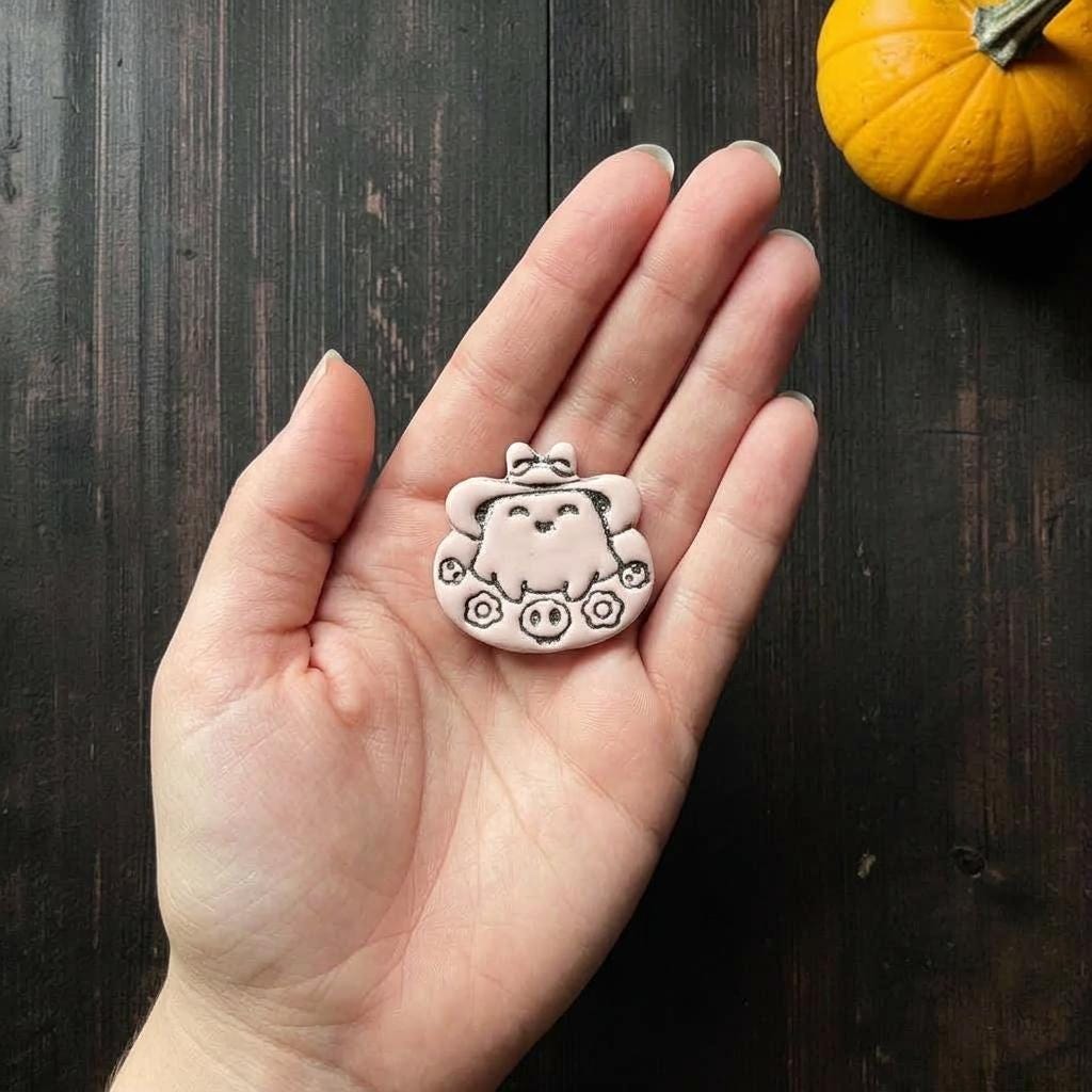 Finished pink and black ghost clay piece resting in a hand for scale, showing the cute ghost character, oversized hat, bow, flowers, and skull details.