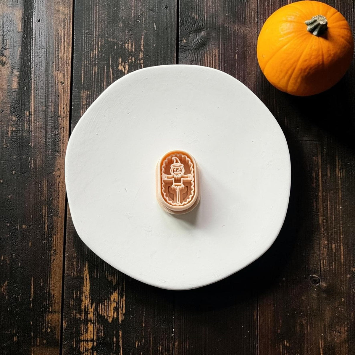 Orange pumpkin scarecrow polymer clay cutter centered on a large white round surface against dark rustic wood, with an orange pumpkin in the upper right corner.