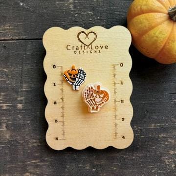 mall finished orange jack-o-lantern held by a white skeleton hand displayed beside the matching cutter on a wooden Craft Love Designs measurement card, with a pumpkin in the background.