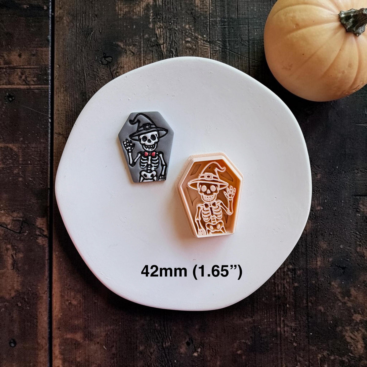 Finished gray coffin-shaped clay piece with a white skeleton wearing a witch hat beside the matching peach-colored design, with text showing 42mm (1.65").