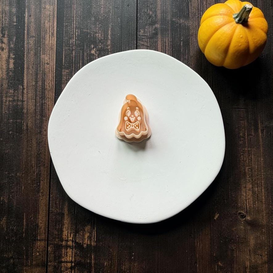 Peach-colored vintage ghost polymer clay cutter featuring a smiling retro face and bow tie, centered on a white round surface with an orange pumpkin at the upper right.