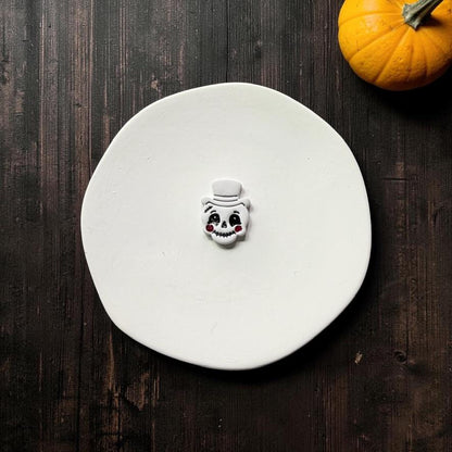 Finished white vintage-style jack-o-lantern face with a top hat, black facial details, toothy grin, and dark red cheeks on a white round surface with an orange pumpkin nearby.