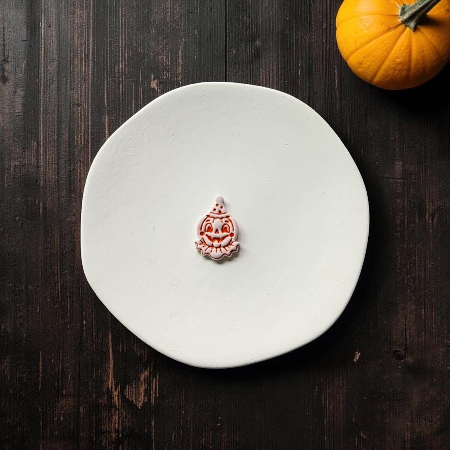 Finished light-colored vintage clown jack-o-lantern clay piece with orange facial details, pointed hat, wide pumpkin grin, and ruffled collar on a white round surface.