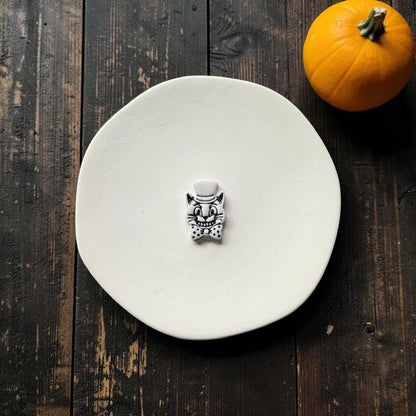 Finished white and black vintage Halloween cat with pointed ears, top hat, wide smiling face, whiskers, and oversized bow tie, centered on a white round surface with an orange pumpkin nearby.