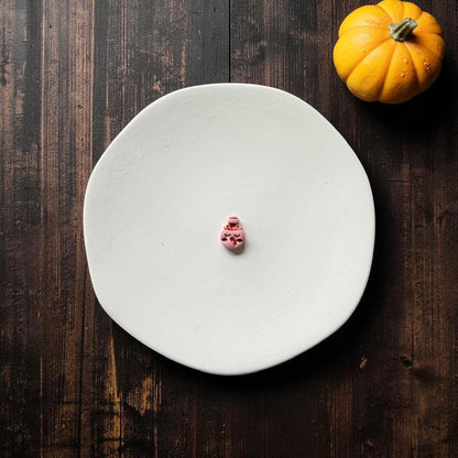 Finished pink turkey character wearing a small pilgrim-style hat with dark facial and feather details, centered on a white round surface with an orange pumpkin in the upper right.