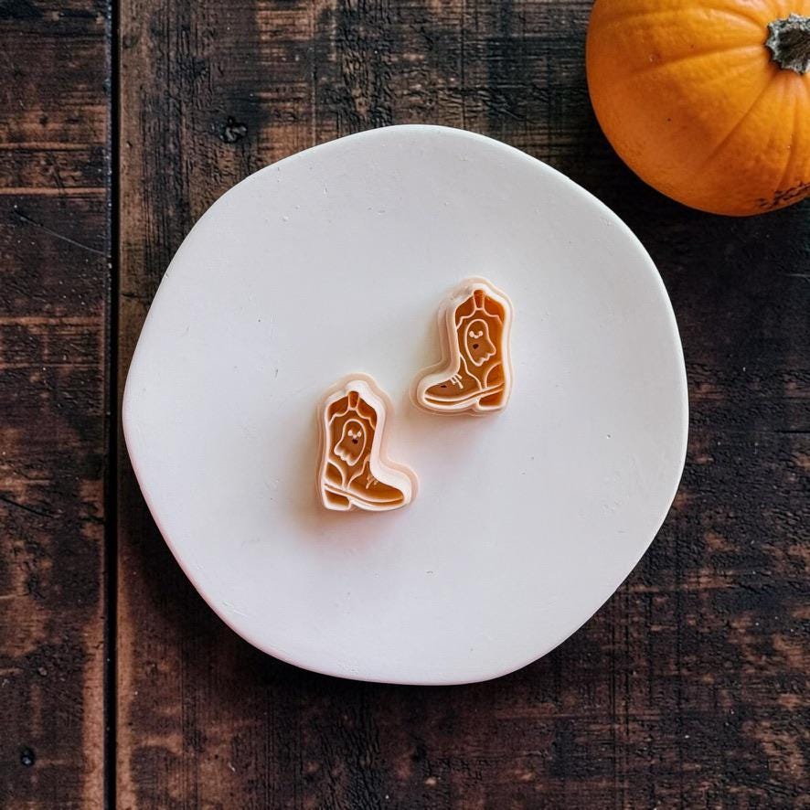 Two peach-colored mirrored cowboy boot cutters featuring small ghost designs displayed on a white round surface with an orange pumpkin.