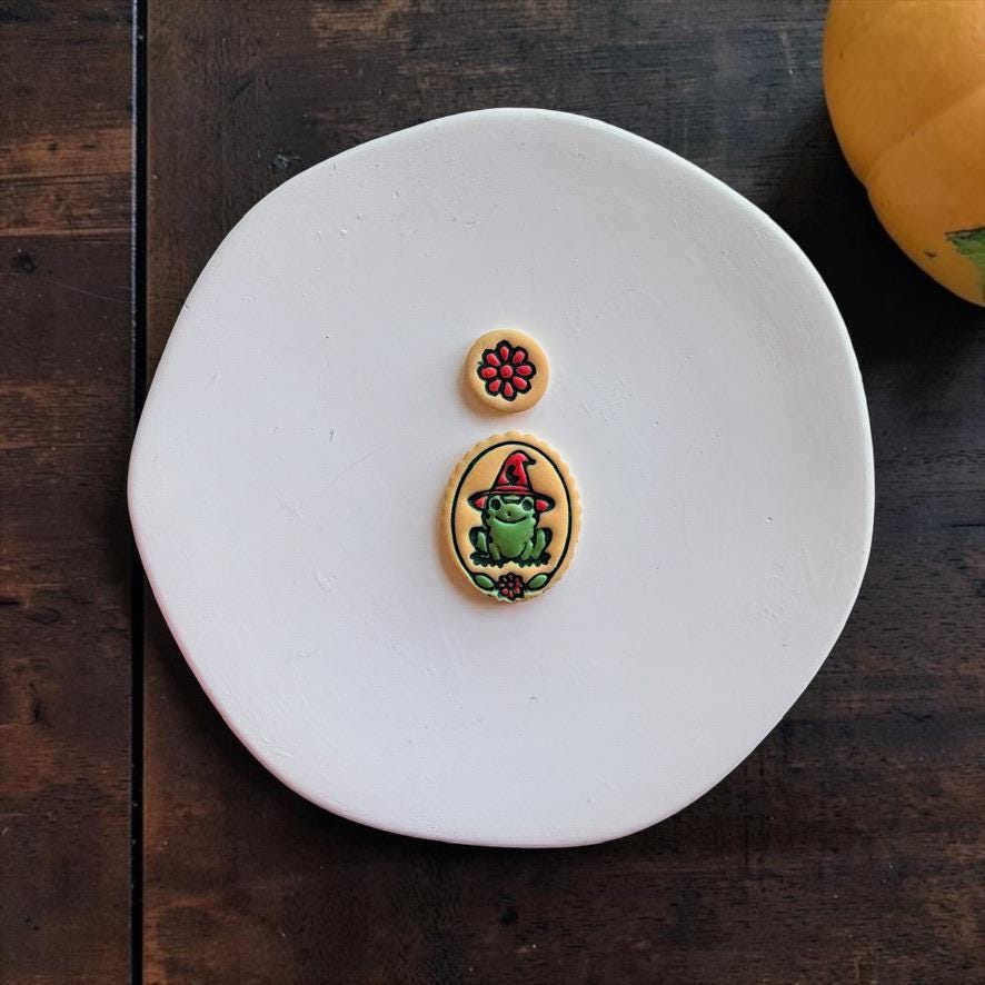Finished witchy frog and flower clay pieces displayed on a white round surface. The green frog wears a red pointed witch hat inside an oval frame with a red flower below, paired with a separate red floral piece.