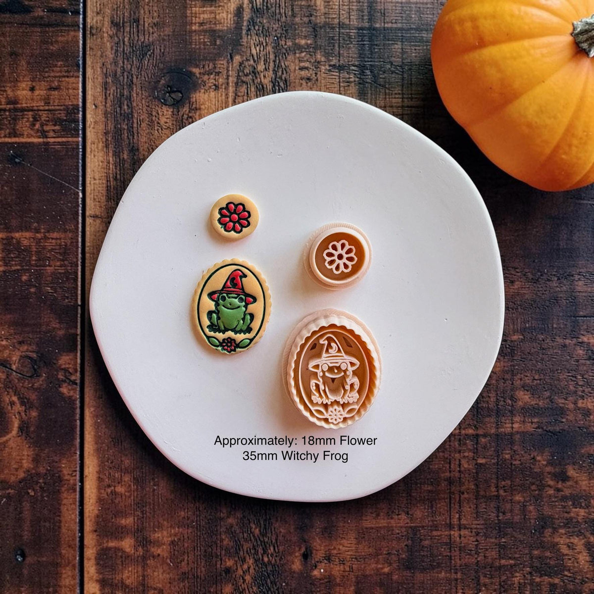 Finished colorful witchy frog and red flower clay pieces displayed beside their matching peach-colored designs on a white round surface with an orange pumpkin. Text states approximately 18mm flower and 35mm witchy frog.