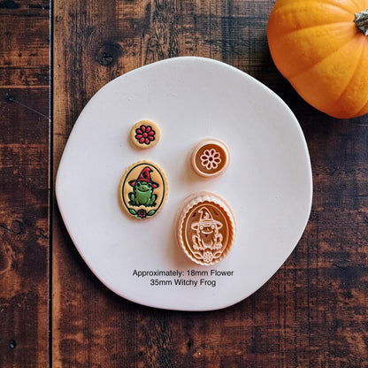 Finished colorful witchy frog and red flower clay pieces displayed beside their matching peach-colored designs on a white round surface with an orange pumpkin. Text states approximately 18mm flower and 35mm witchy frog.