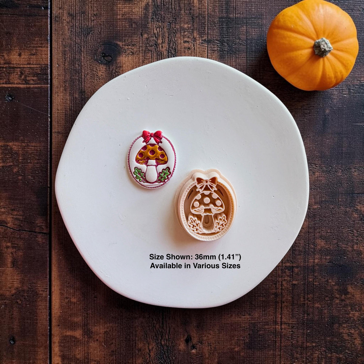 Finished colorful mushroom piece displayed beside the matching peach-colored design on a white round surface. The mushroom has a yellow and red spotted cap, green leaves, red bow, and oval border. Text states size shown 36mm (1.41 inches) and available in various sizes.