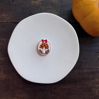 Finished colorful oval mushroom design centered on a white round surface against a dark wood background, with an orange pumpkin visible in the upper right corner.