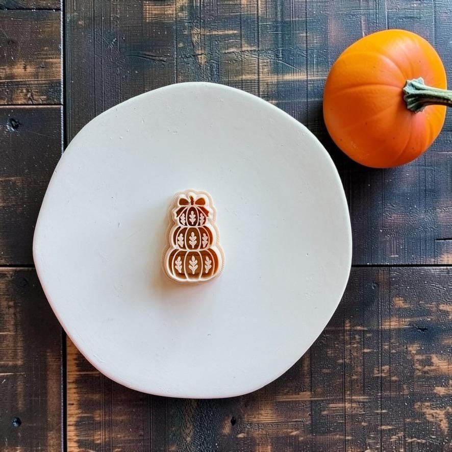 Peach-colored stacked pumpkin design centered on a white round surface with an orange pumpkin in the upper right. The design features three decorated pumpkins topped with a bow.