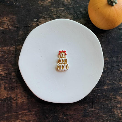 Finished white, gold, and red stacked pumpkin clay piece centered on a white round surface against a dark rustic wood background, with an orange pumpkin in the upper right.