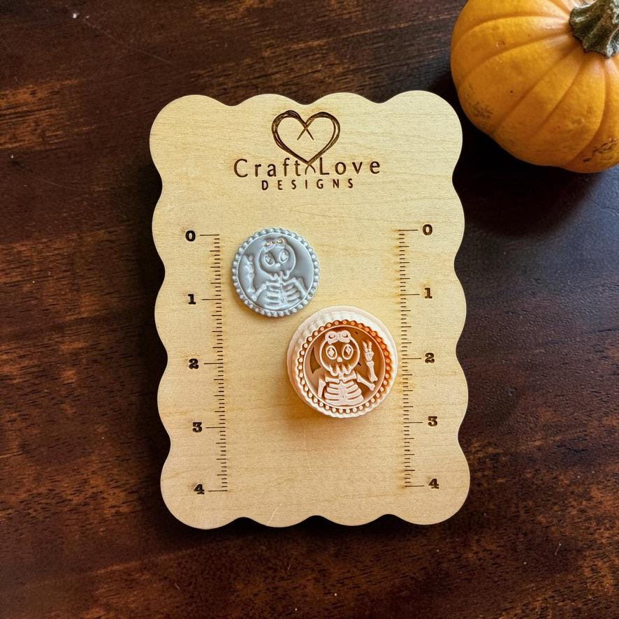 Gray finished clay skeleton girl with one hand making a peace sign beside a larger peach-colored version on a Craft Love Designs wooden measuring board.