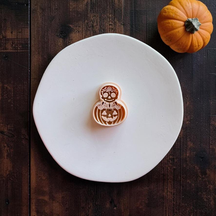 Peach-colored skeleton and jack-o-lantern design centered on a white round surface, showing a decorated skull above a smiling carved pumpkin.
