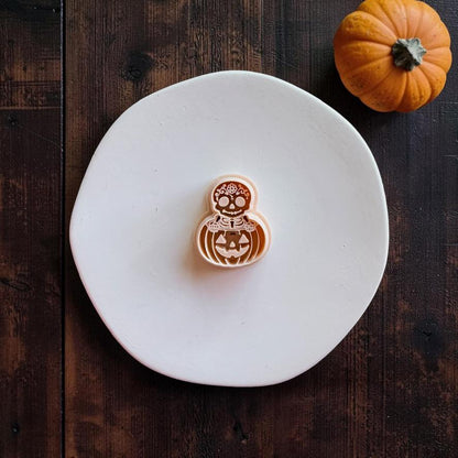 Peach-colored skeleton and jack-o-lantern design centered on a white round surface, showing a decorated skull above a smiling carved pumpkin.