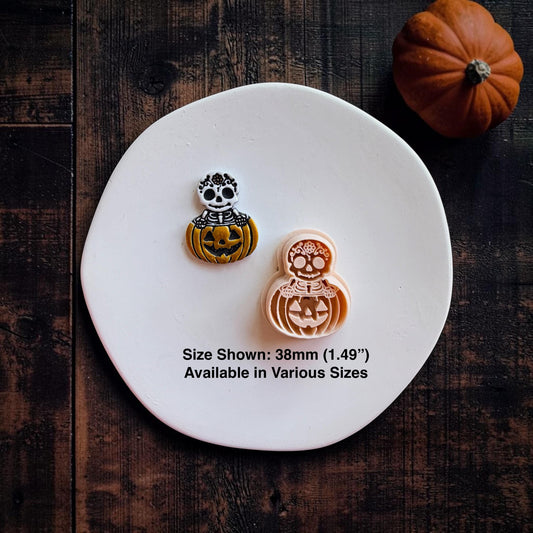 Finished black, white, and orange skeleton pumpkin clay piece beside the peach-colored version on a white round surface. Text states size shown is 38mm and available in various sizes.
