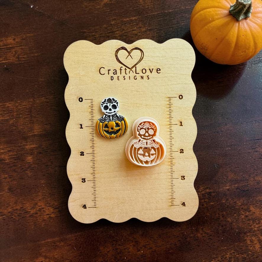 Finished skeleton jack-o-lantern example displayed beside the peach-colored version on a Craft Love Designs wooden measuring board, with an orange pumpkin in the background.