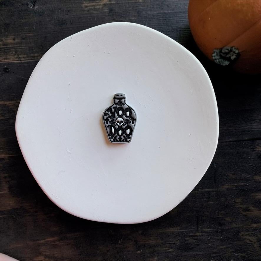Black and gray finished potion bottle centered on a white round surface, featuring a small white skull surrounded by dark decorative lettering and ornate flourishes.
