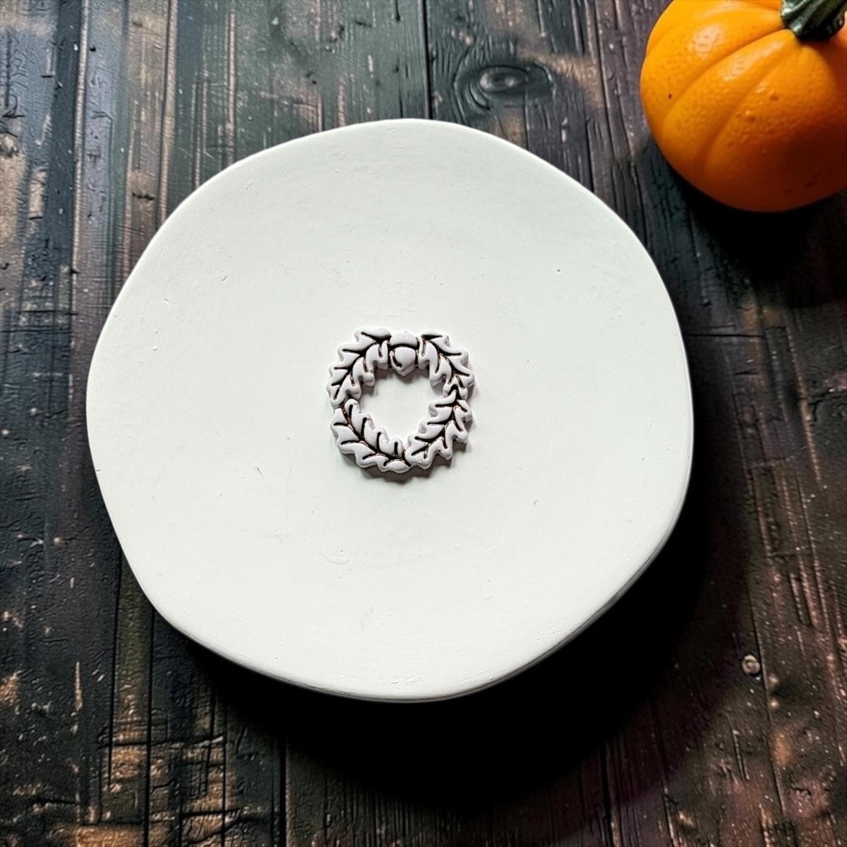 Finished white clay acorn and oak leaf wreath with dark outlined leaf details centered on a white round surface, with an orange pumpkin in the upper right.