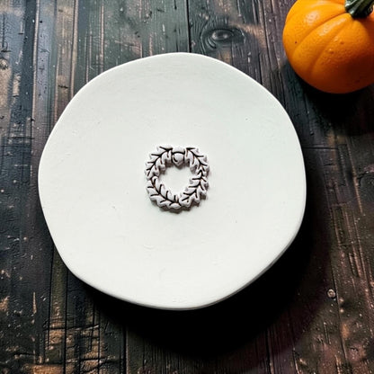 Finished white clay acorn and oak leaf wreath with dark outlined leaf details centered on a white round surface, with an orange pumpkin in the upper right.