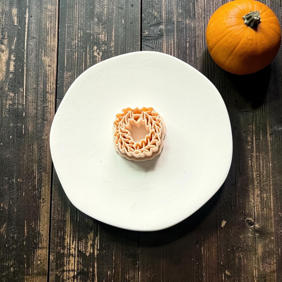 Orange acorn and oak leaf wreath polymer clay cutter centered on a white round surface against a dark rustic wood background with an orange pumpkin.