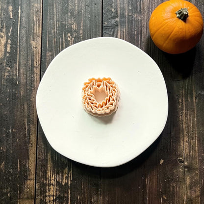 Orange acorn and oak leaf wreath polymer clay cutter centered on a white round surface against a dark rustic wood background with an orange pumpkin.
