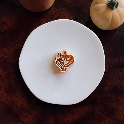 Peach-colored pumpkin and skeleton hand polymer clay cutter centered on a white round surface against a dark wooden background with pumpkins nearby.