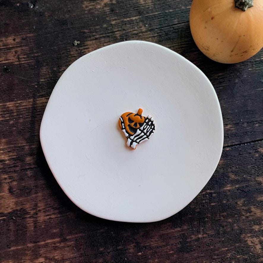 Finished orange jack-o-lantern wrapped in a white skeleton hand centered on a white round surface against a dark rustic wooden background with a pumpkin in the upper right.